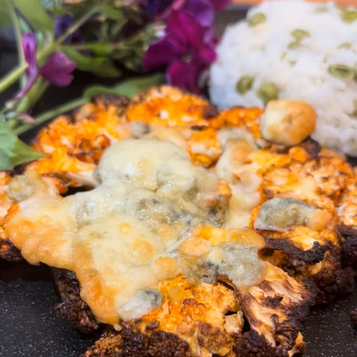 Buffalo Cauliflower Steak on a black square plate with white rice and peas and purple flowers