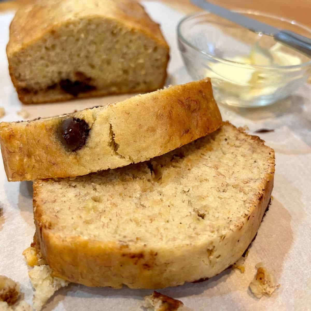 Slices of chocolate chip banana bread on parchment paper with a small glass bowl filled with butter