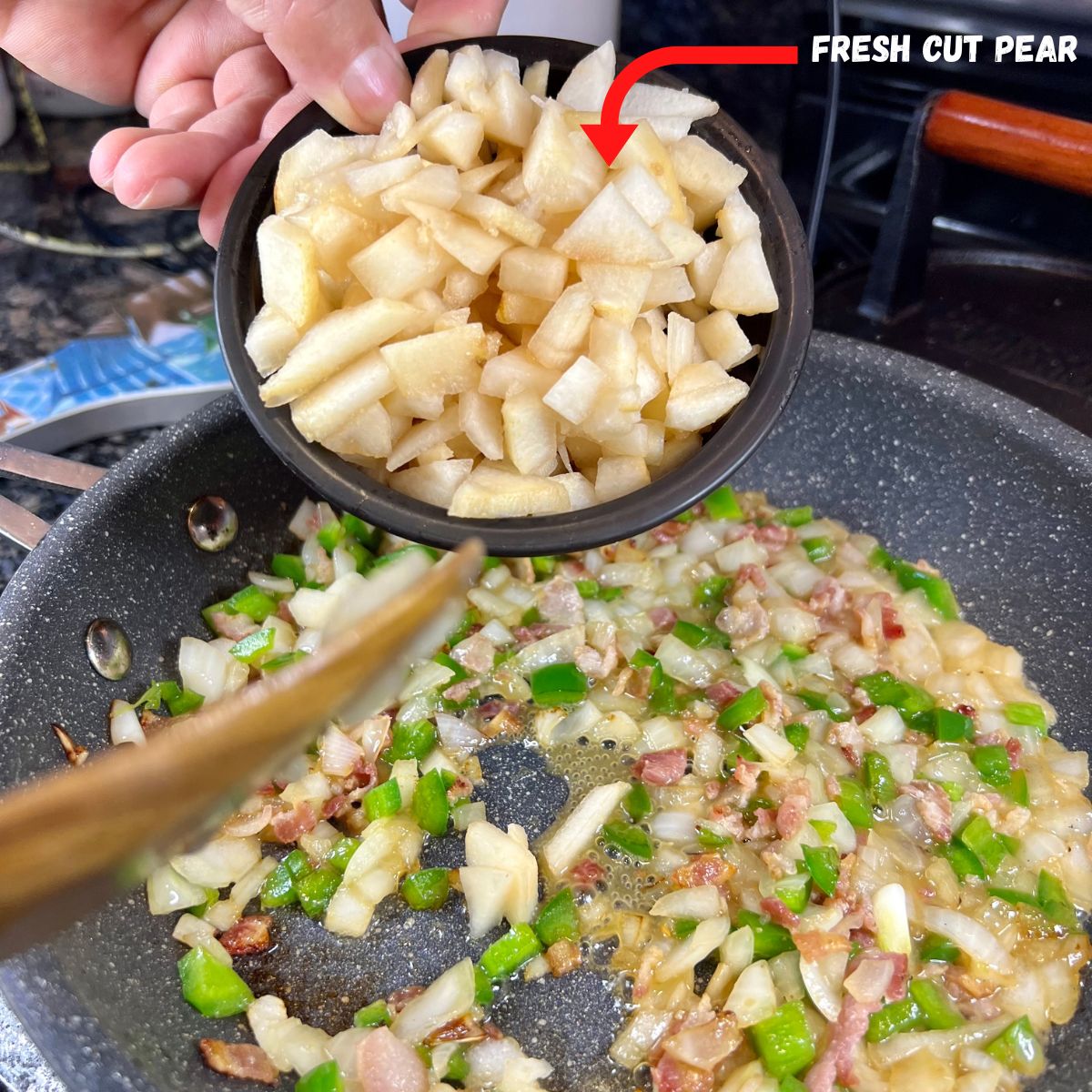 A hand holding a bowl of diced pear over a frying pan with bacon onion an jalapeños cooking