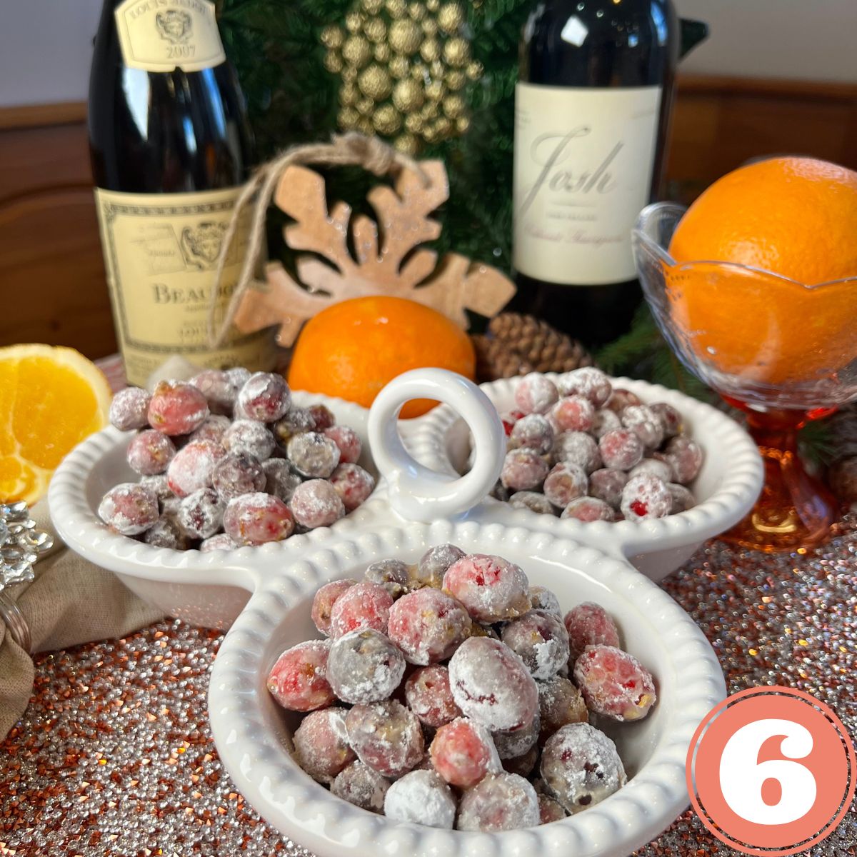 Candied cranberries in a tripe white serving tray on a table with 2 bottles of red wine and 2 oranges