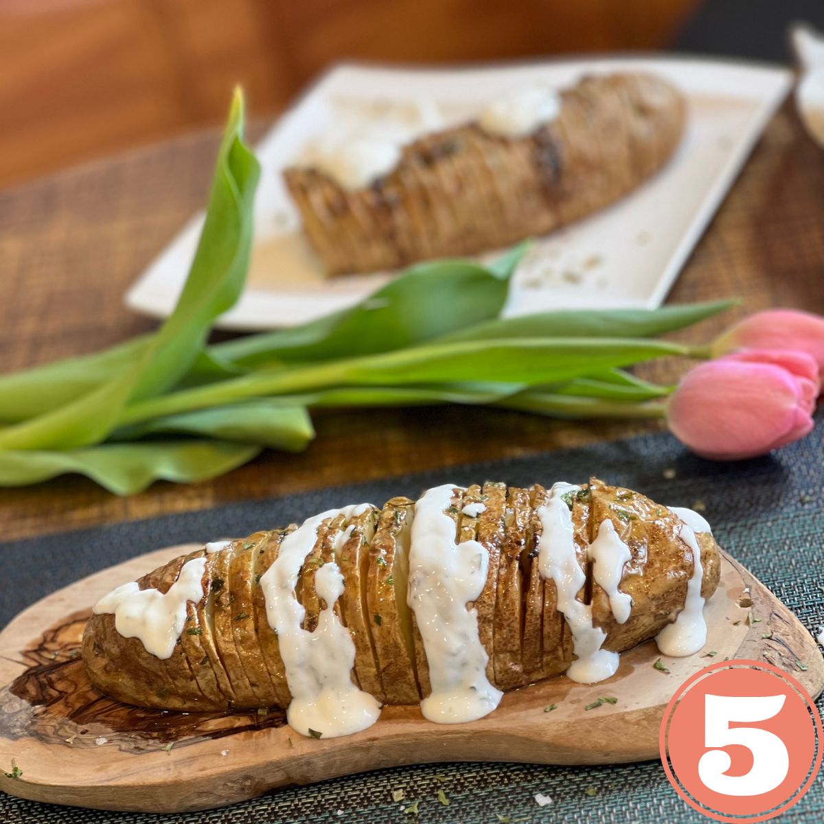 Ranch dressing drizzled over a WW russet hasselback potato on a table with a pink tulip