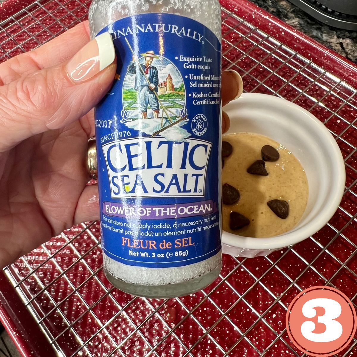 A hand holding a jar of Celtic sea salt over cookie baked oats in a white ramekin on a red baking pan