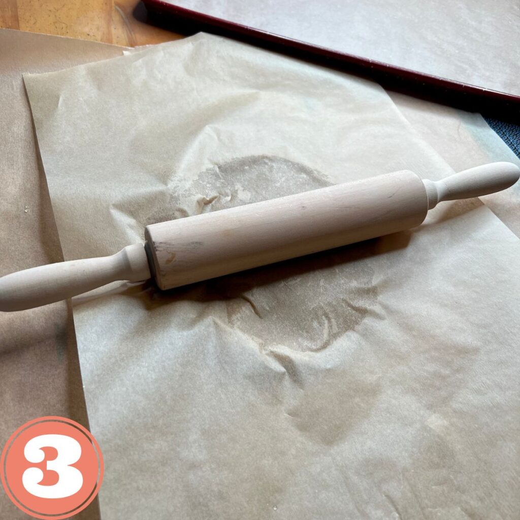 A wooden rolling pin on a sheet of brown parchment paper flattening dough