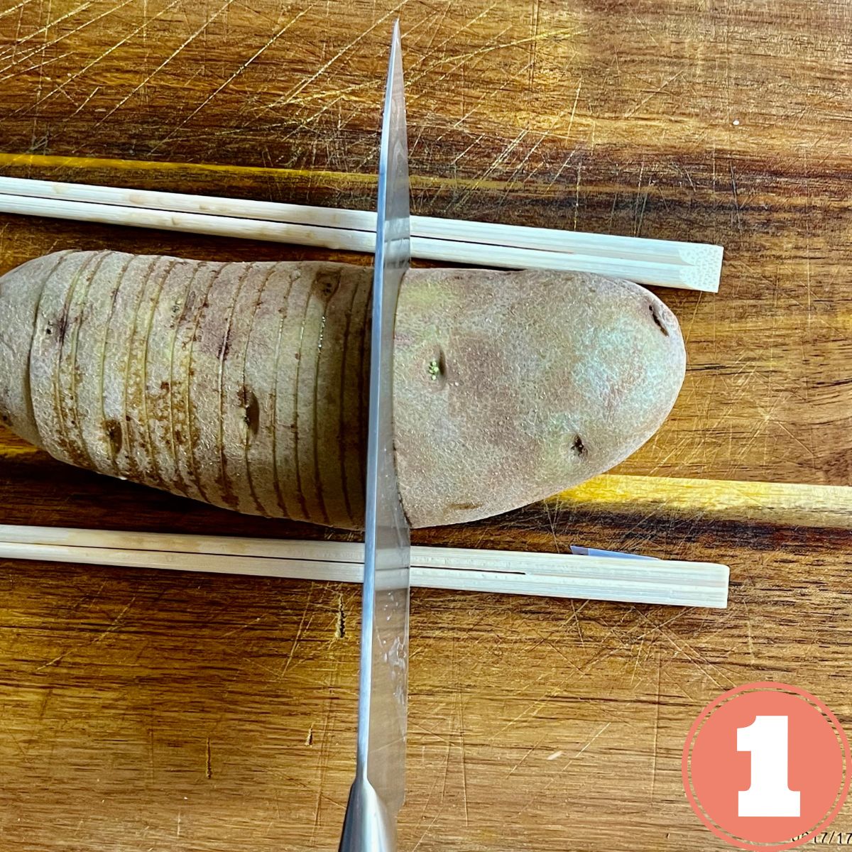 A russet potato on a wooden cutting board with chopsticks supporting the sides being sliced into a hasselback potato