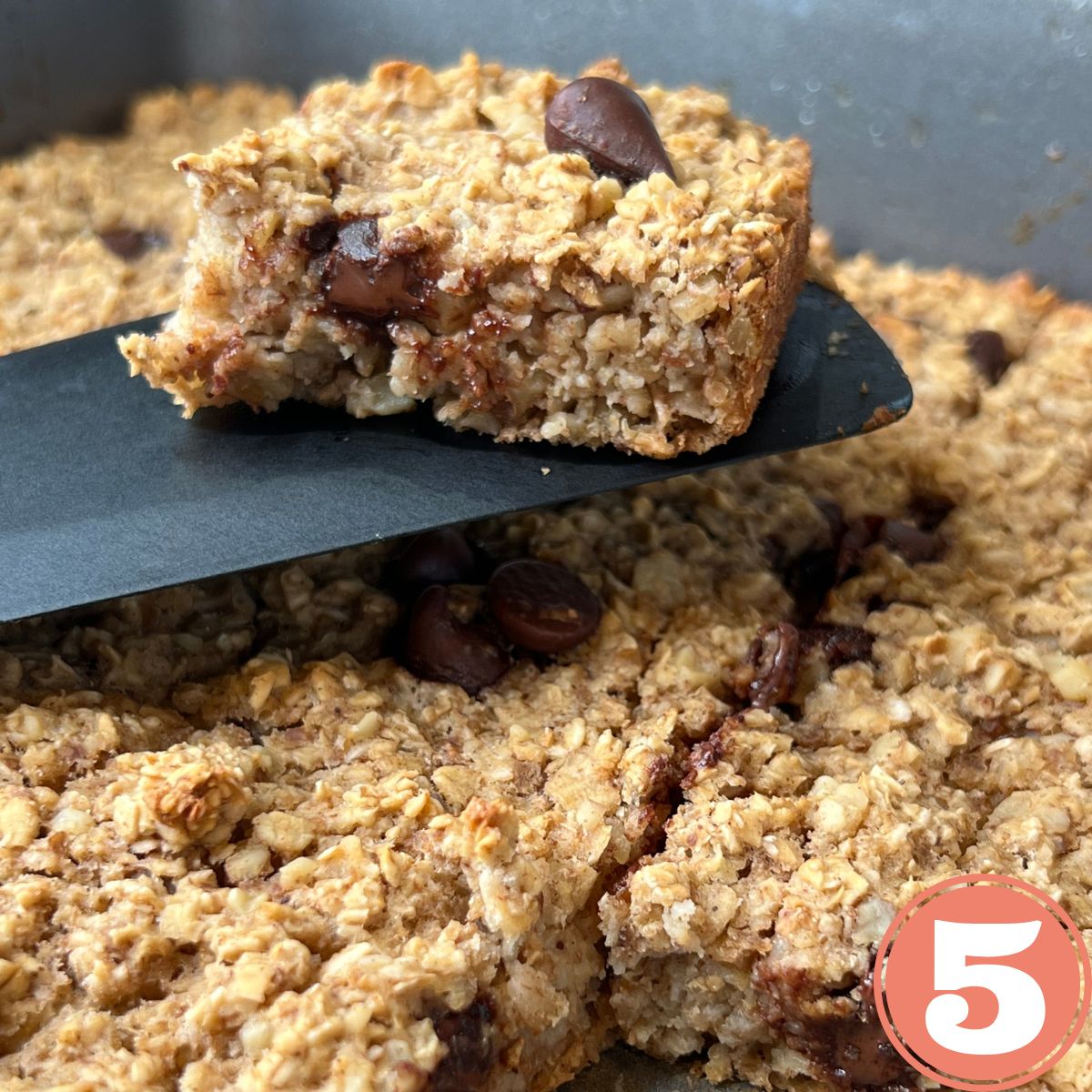 A slice of baked oatmeal with chocolate chips on a black spatula over a pan of baked oatmeal