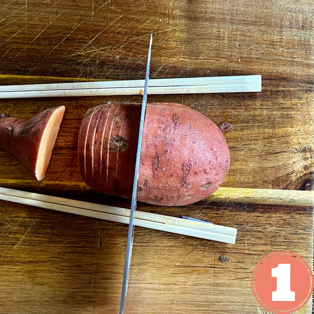A sweet potato bing sliced with a large knife hasselback style on a wooden cutting board with chop sticks