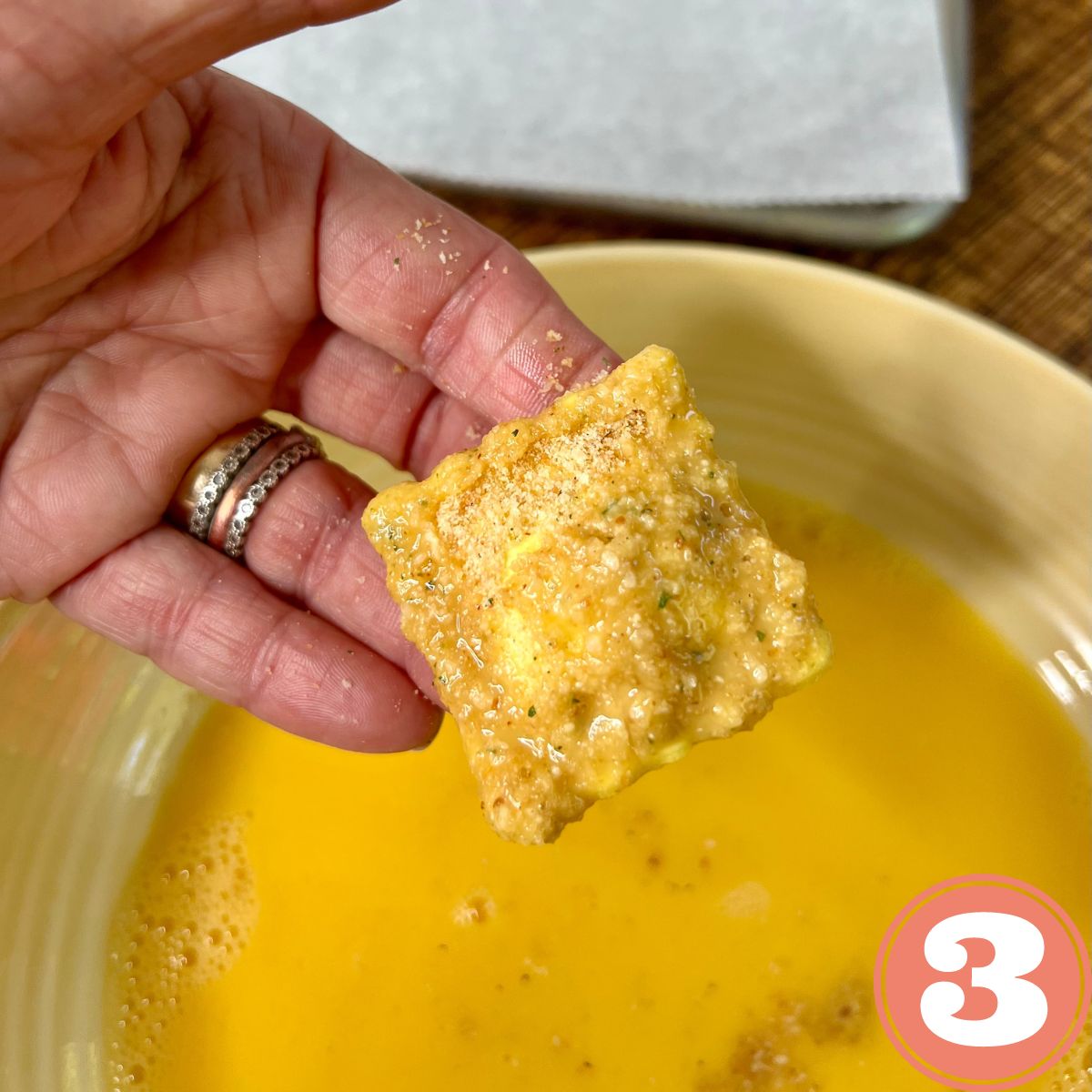 A hand holding a breaded frozen ravioli over a bowl of egg wash