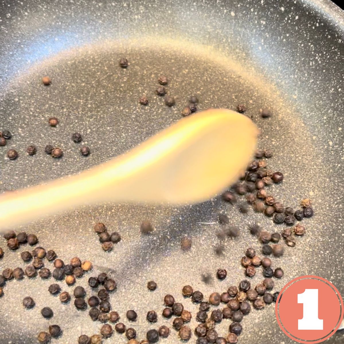 Whole black peppercorns being toasted in a frying pan with a wooden spoon stirring them