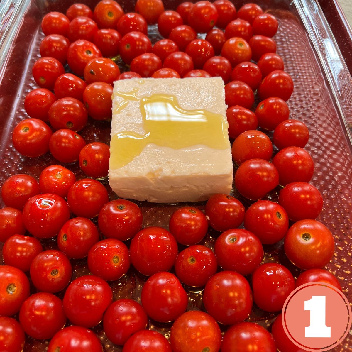 Cherry tomatoes in a glass baking dish with a block of feta cheese drizzled with olive oil