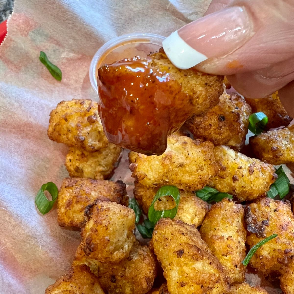 A hand holding an air fried tater tot dipped in hot sauce