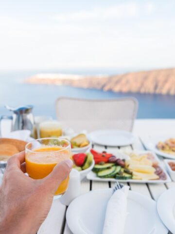 a hand holding a glass of juice over a table of breakfast foods by the ocean