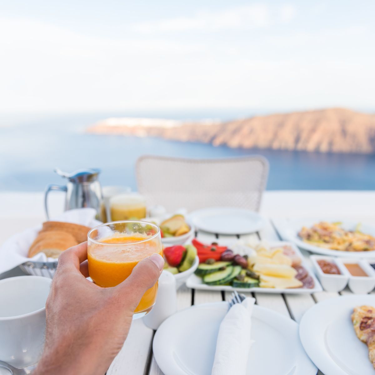 a hand holding a glass of juice over a table of breakfast foods by the ocean
