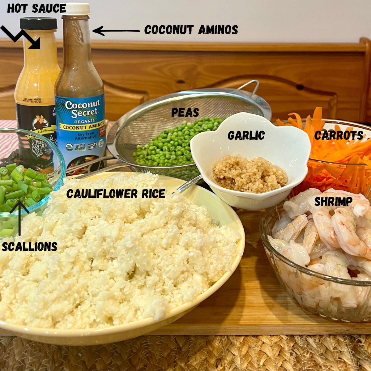 A bowl of riced cauliflower, a bottle of hot sauce and coconut aminos, scallions, garlic, carrots and fresh shrimp
