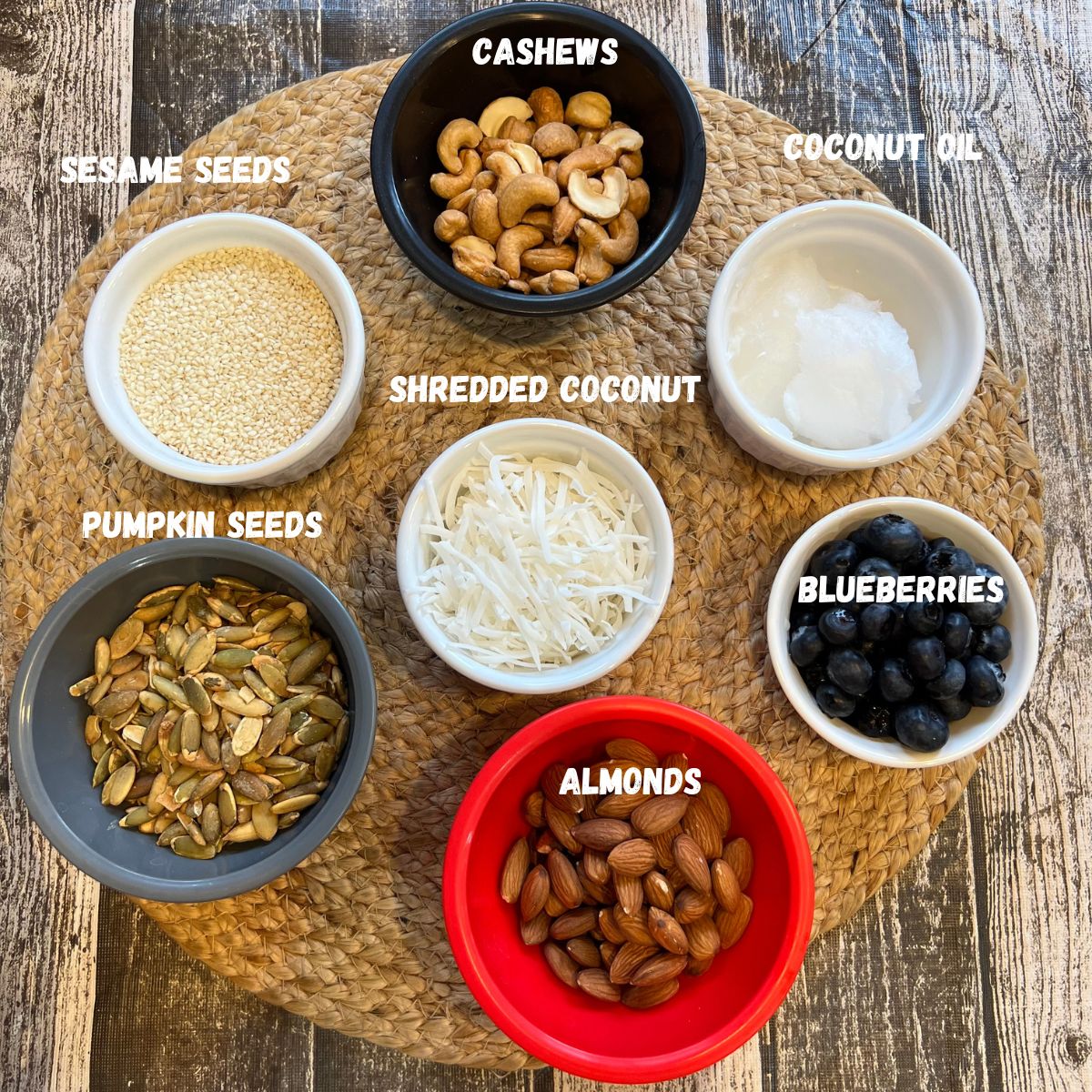 Homemade granola ingredients - sesame seeds, cashews, coconut oil, pumpkin seeds, coconut flakes, blueberries and almonds in small bowls