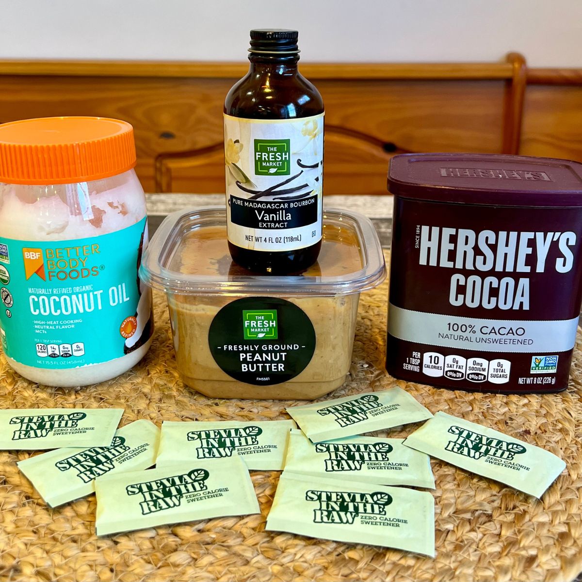 A jar of coconut oil,a bottle of vanilla extract, a container of peanut butter, a container of Hershey's unsweetened cocoa powder and packets of Stevia on a table