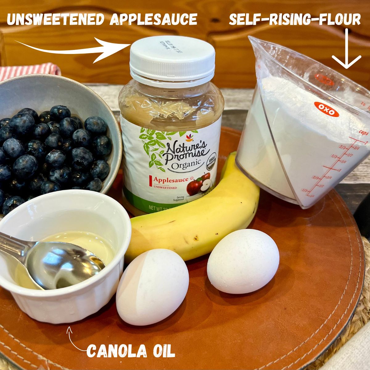 A bowl of blueberries, a jar of applesauce, a measuring cup filled with self-rising flour, a banana, 2 eggs and a bowl of canola oil