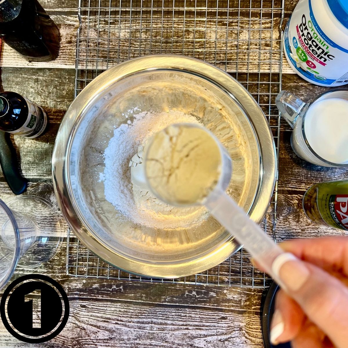 A bowl with Krusteaz Buttermilk Pancake Mix with a hand holding a scoop of protein powder