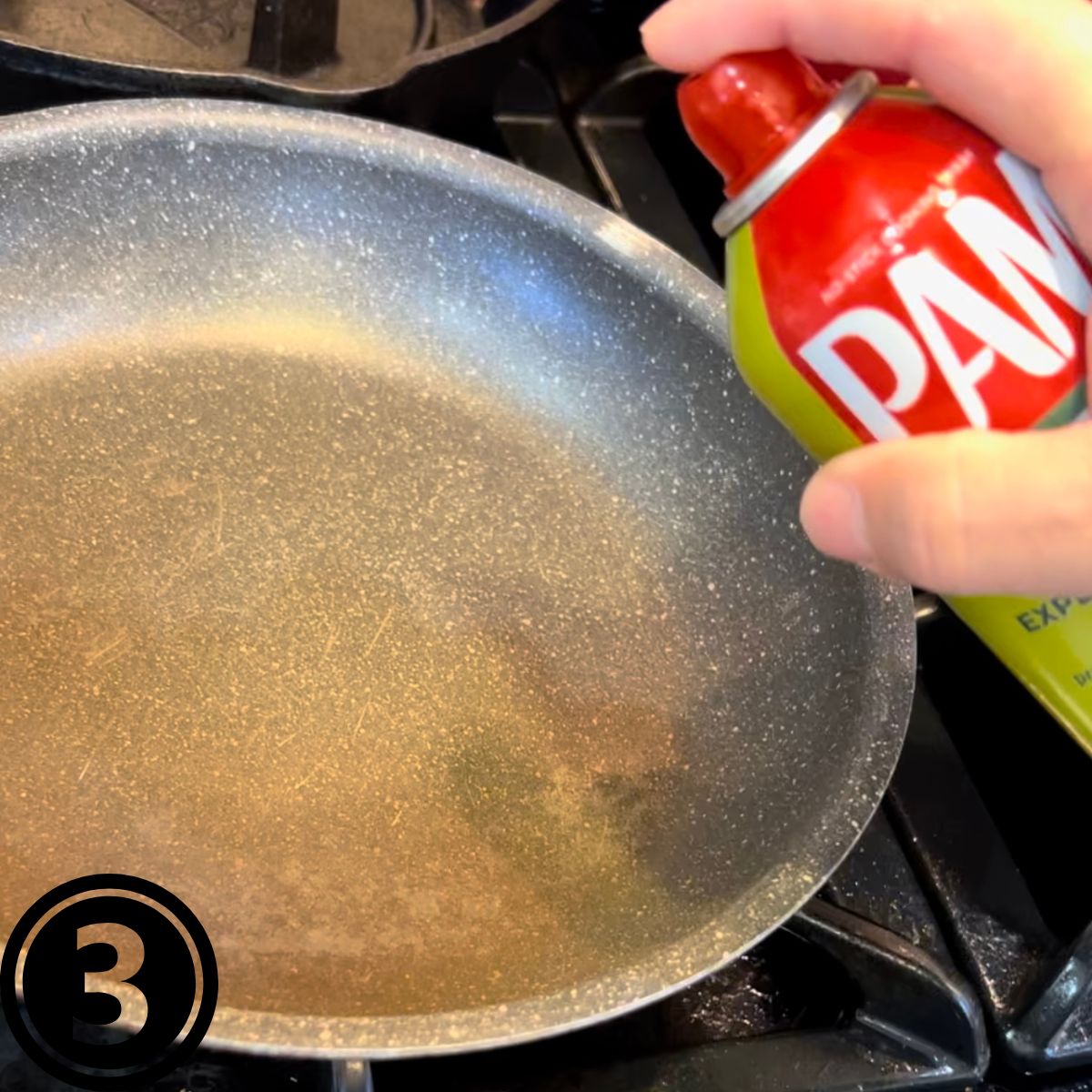 A hand holding a can of PAM spray olive oil over a frying pan