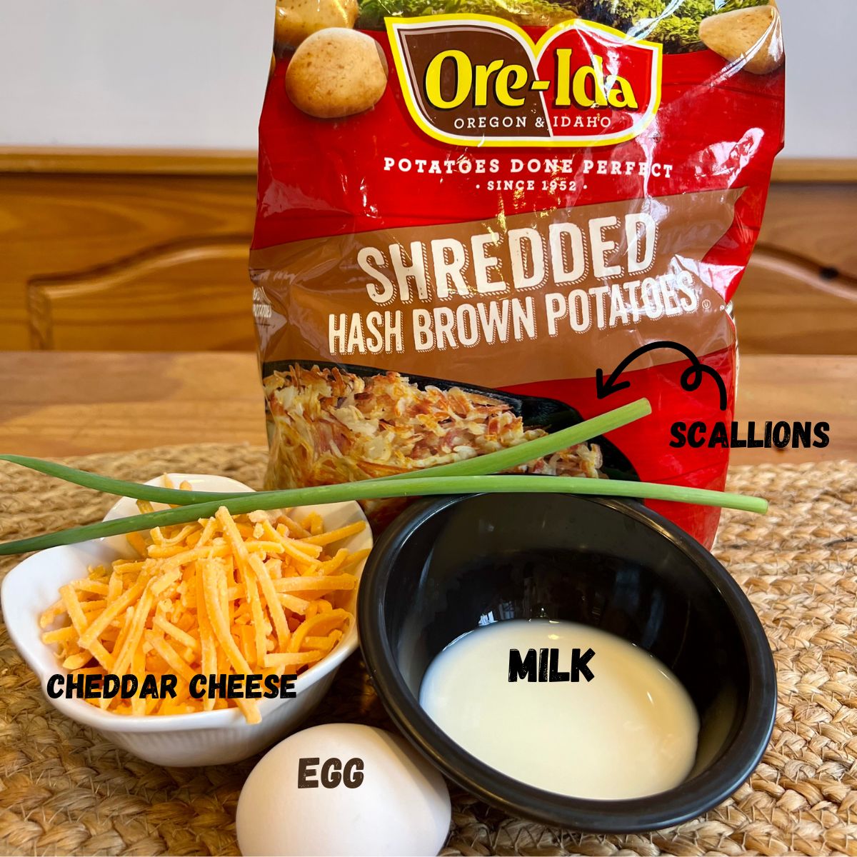 A bowl of shredded cheddar cheese, a bag of Ore-Ida frozen hash browns, 2 scallions, a bowl of milk and an egg on a table