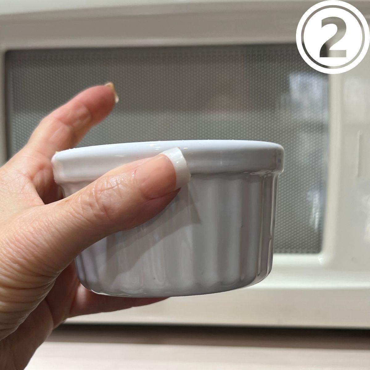 Hand holding a white ramekin in font of a microwave