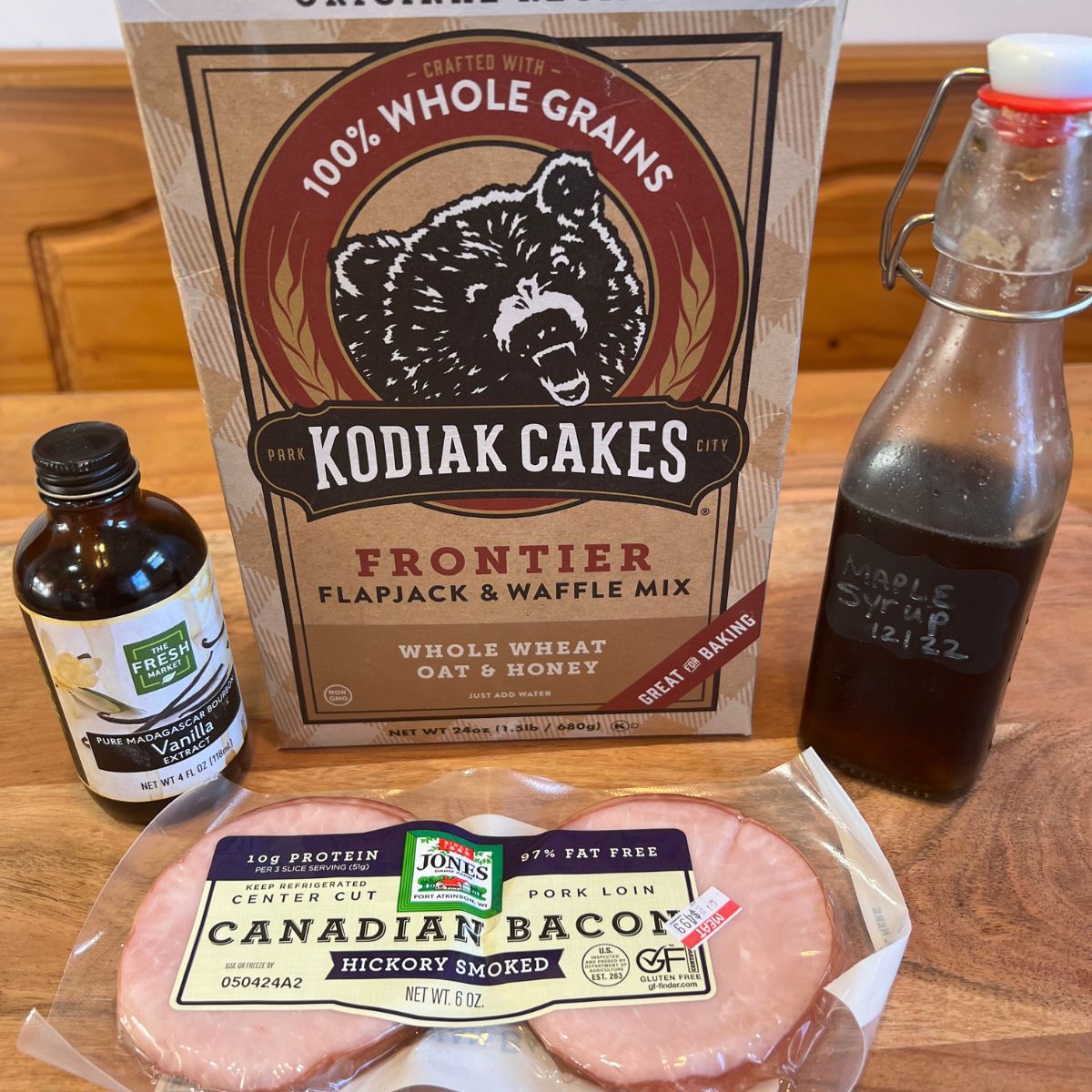 A package of Canadian Bacon and Kodiak Cakes Pancake Mix and maple syrup and vanilla extract on a table