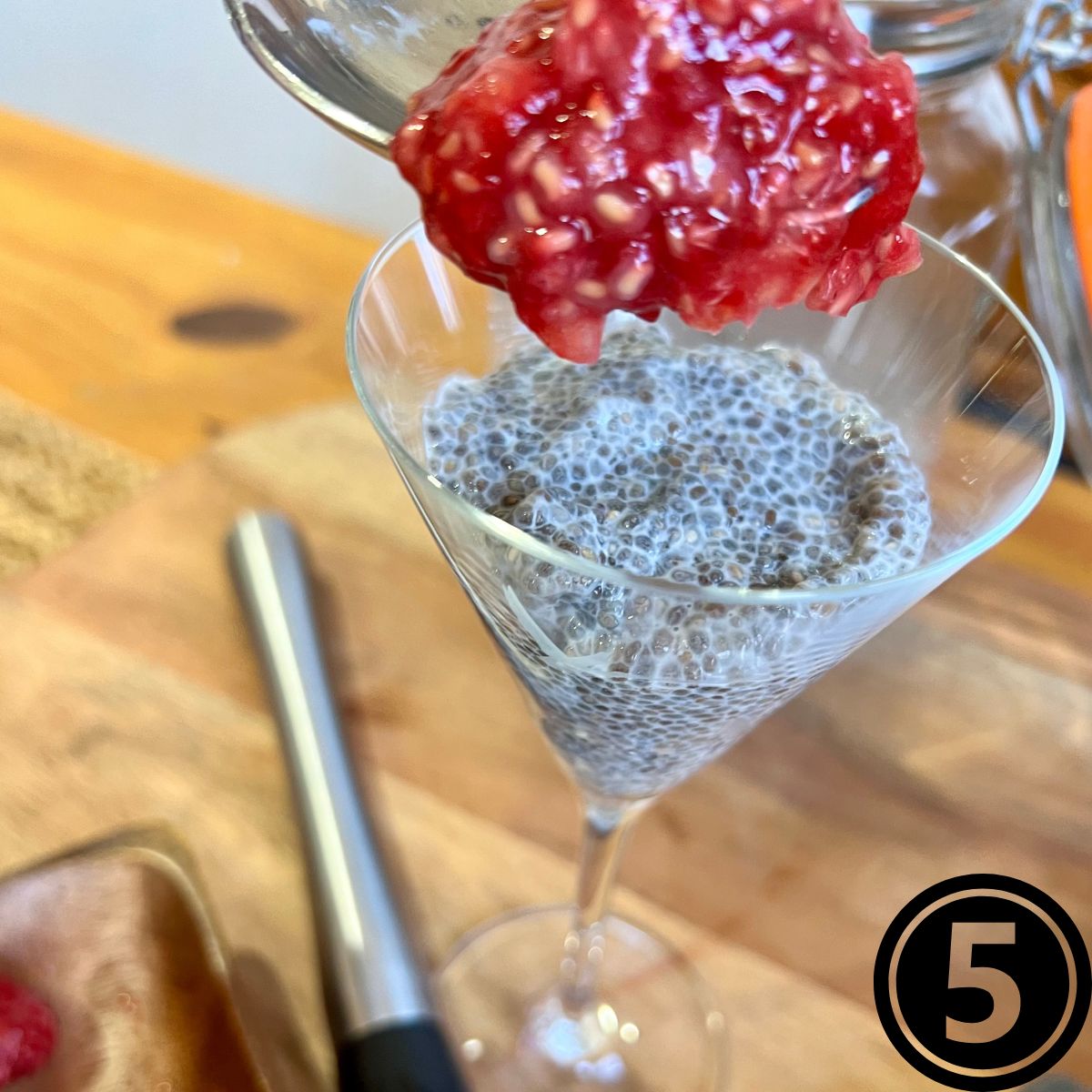 A spoon filled with mashed raspberries being poured on top of a glass a vanilla chia seed pudding