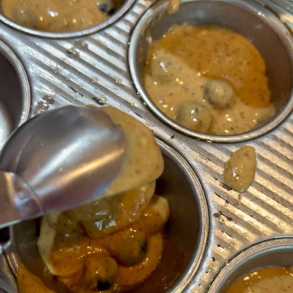 A cookie scoop filling muffin cups with to die for blueberry muffin batter