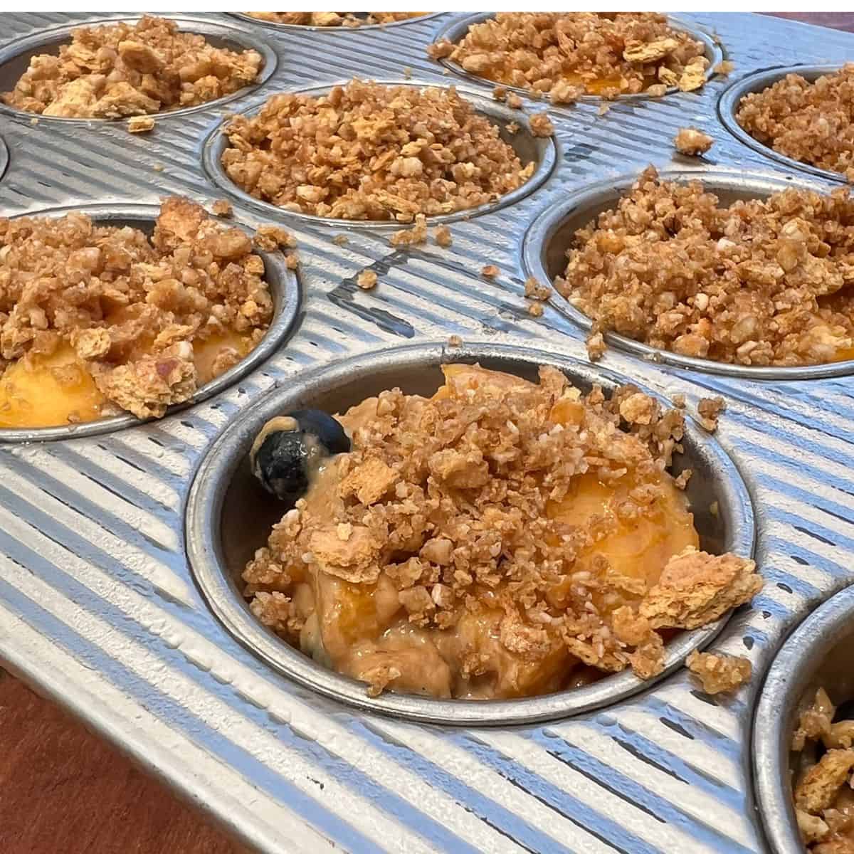Peach blueberry muffin batter covered in graham cracker crumble in muffin cups