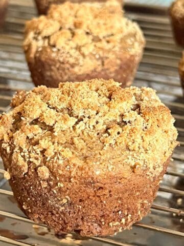 Close-up of a golden brown coffee cake muffin with crumb topping on a wire rack, showing a soft, moist texture and cinnamon streusel top