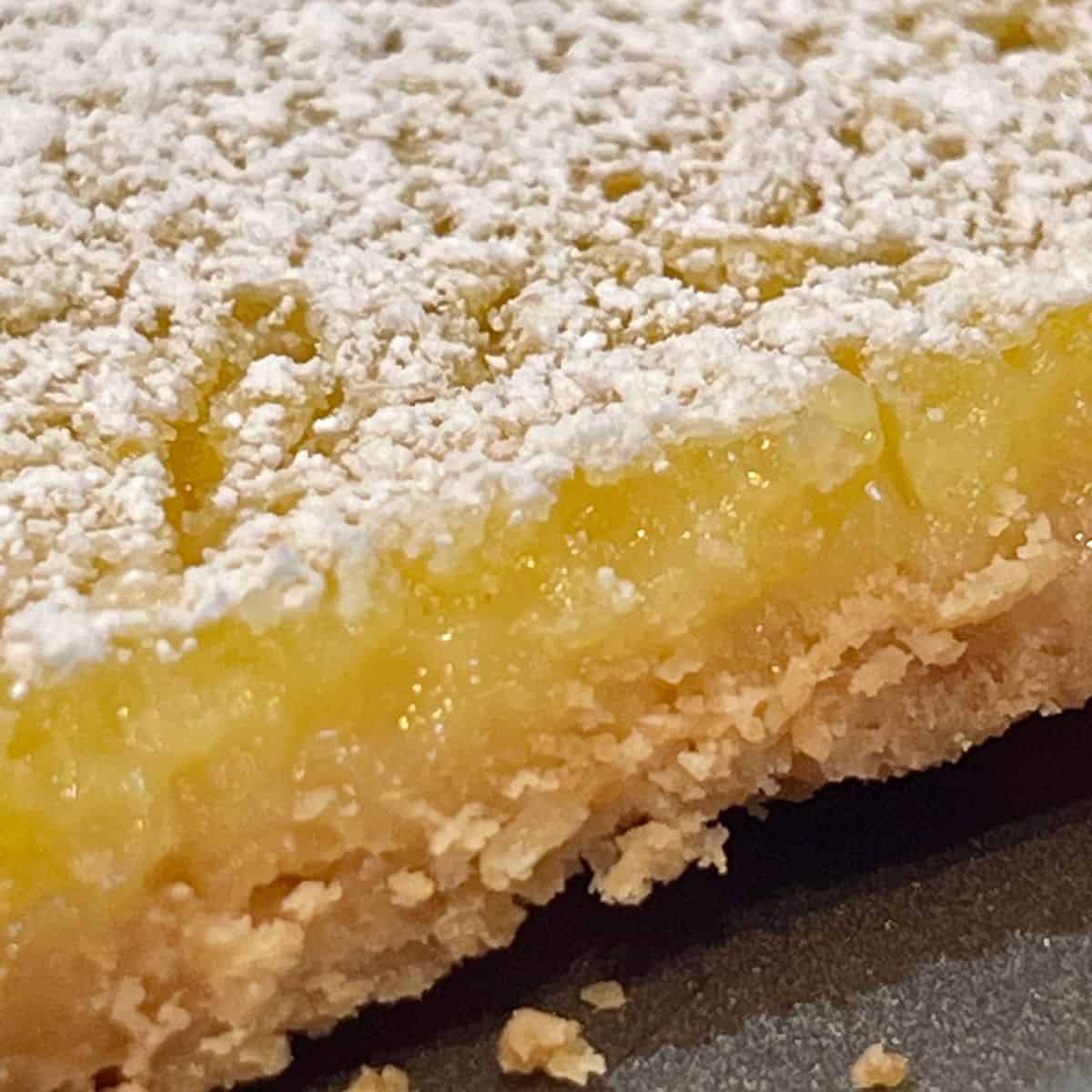 Close up of lemon bar showing buttery crust, creamy lemon filling, and powdered sugar topping