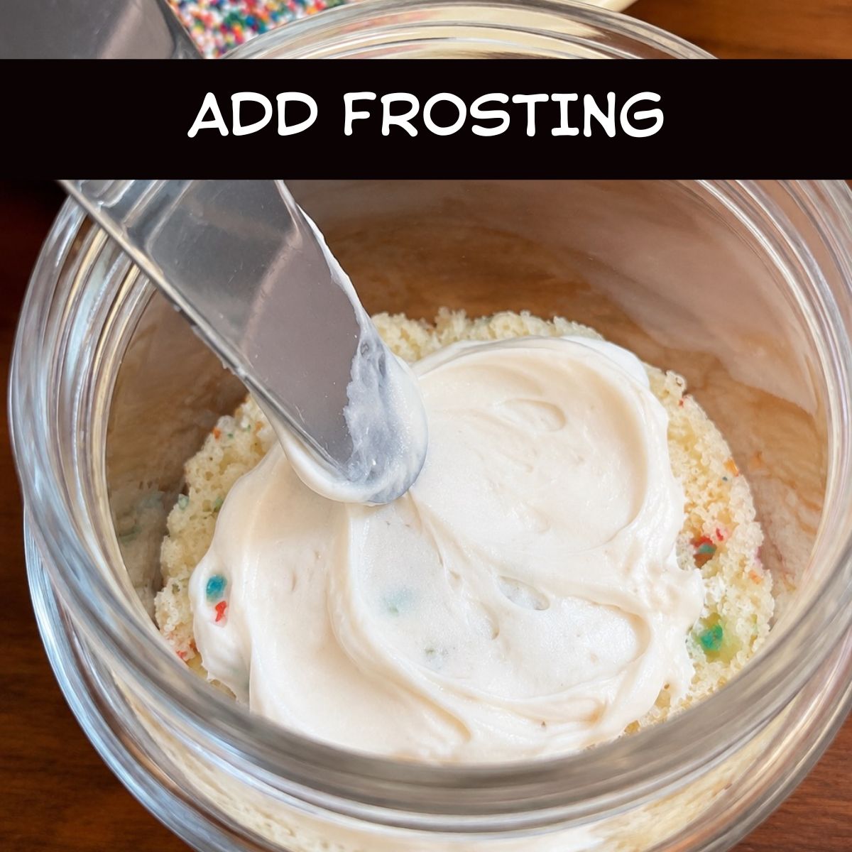 Cream cheese frosting being spread over the first layer of Funfetti cake inside a glass jar while assembling Healthy Funfetti Dot Cake Jars.