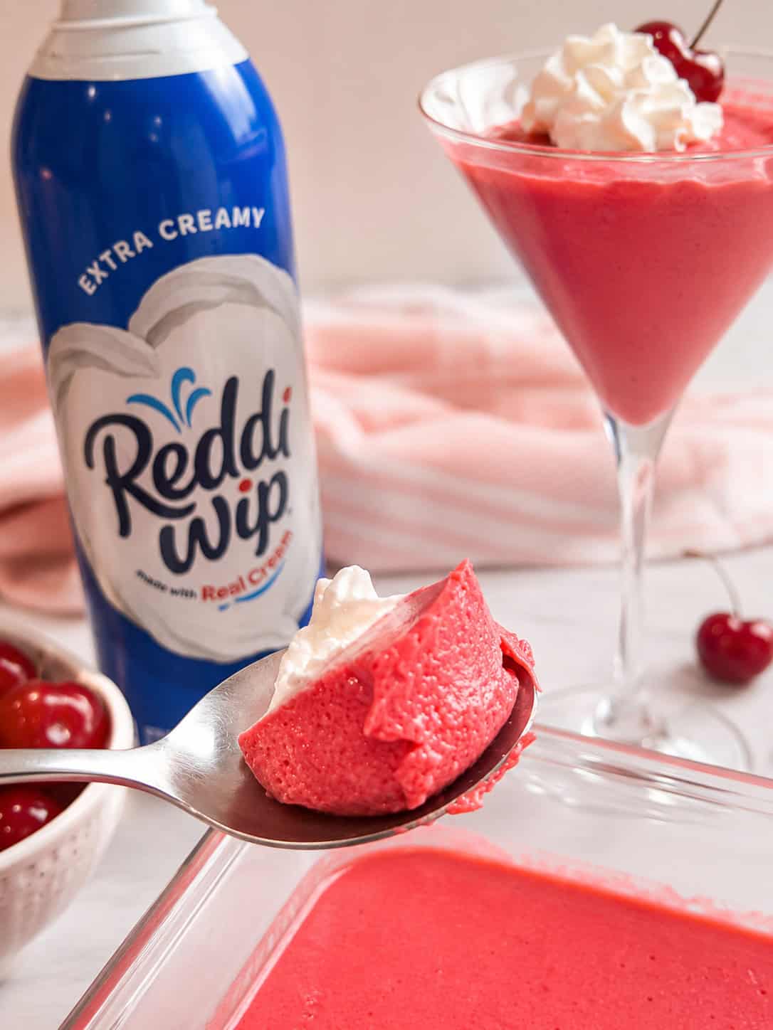 Spoonful of high-protein cherry Jell-O fluff topped with whipped cream, with a serving of the dessert in a martini glass in the background.