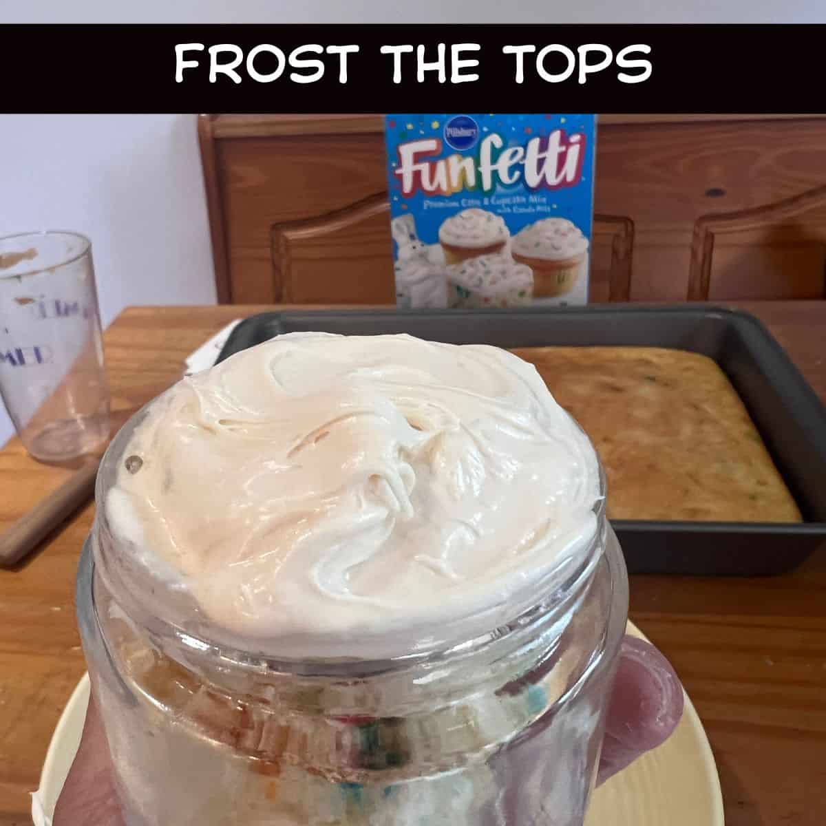 Hand holding a glass jar filled with layered Funfetti cake and cream cheese frosting, topped with a smooth layer of frosting before adding sprinkles.