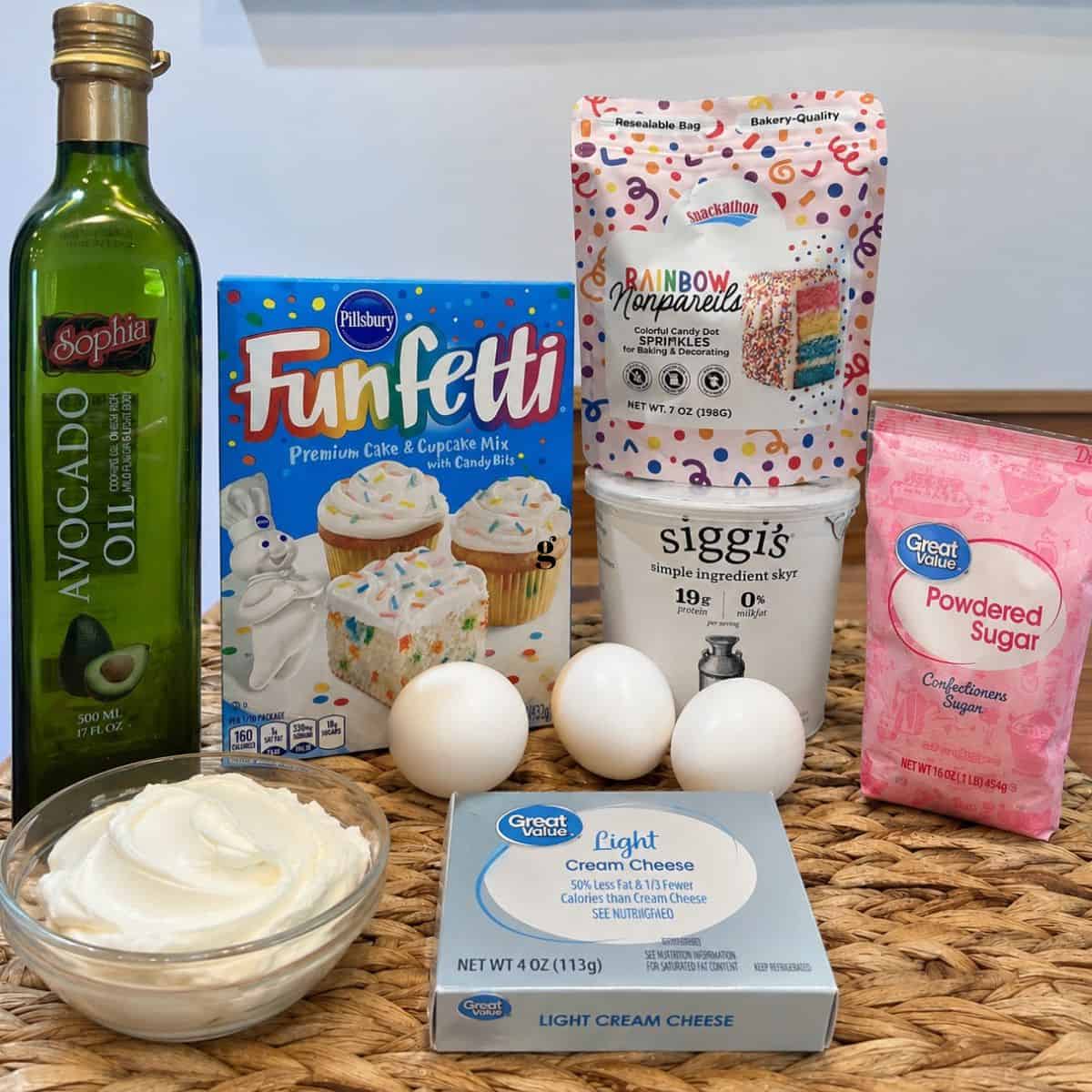 Ingredients for Healthy Funfetti Dot Cake Jars arranged on a tabletop, including Funfetti cake mix, avocado oil, Greek yogurt, light cream cheese, powdered sugar, rainbow nonpareil sprinkles, and eggs.