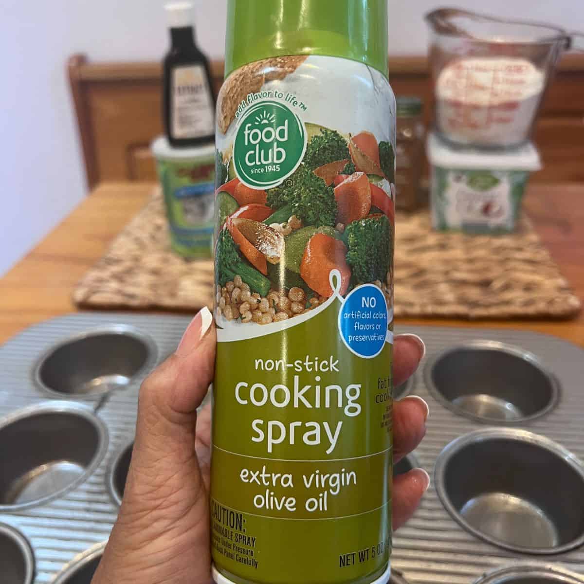 Hand holding olive oil cooking spray over a muffin pan before baking