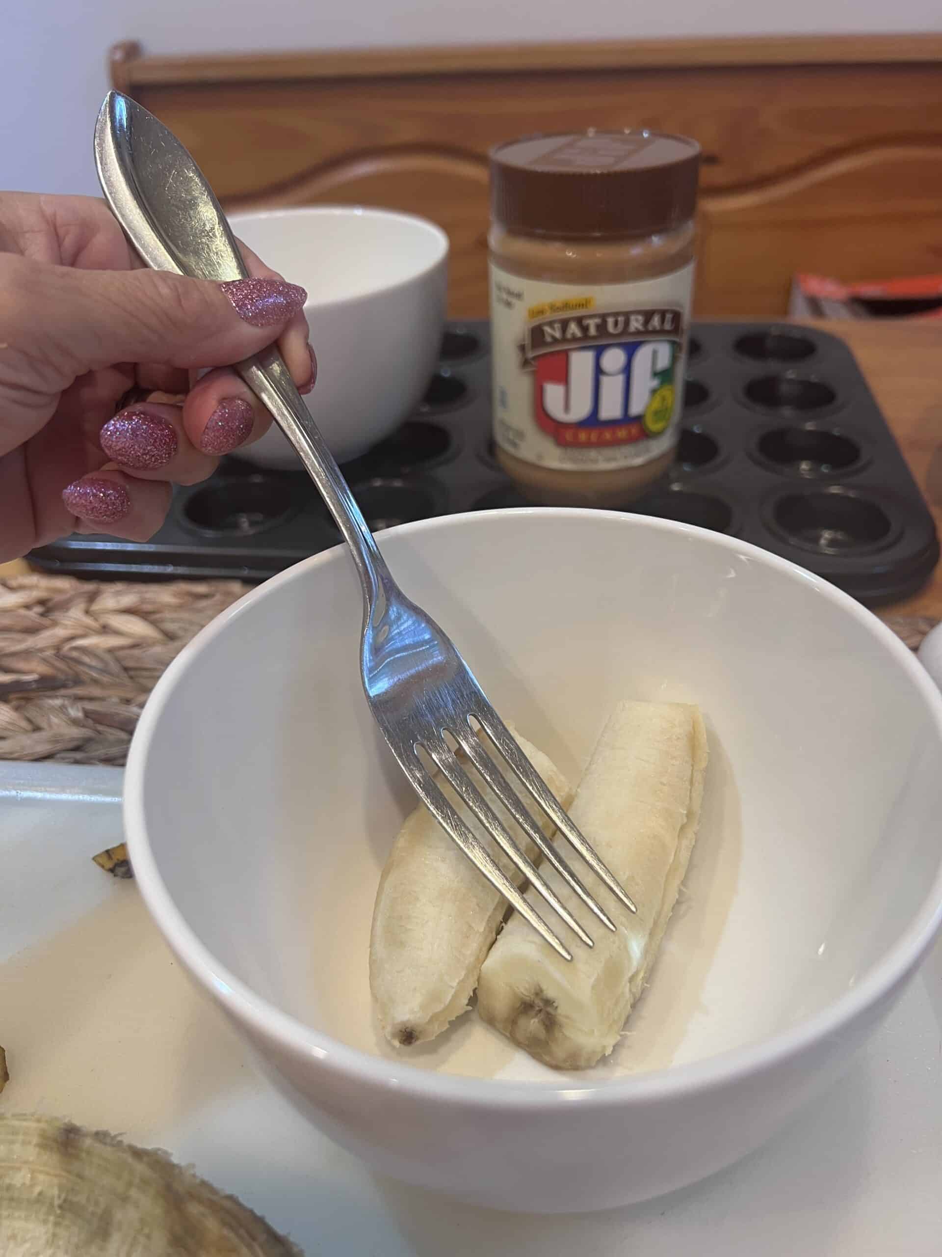 Hand holding a fork over a ripe banana in a mixing bowl, ready to mash it for 3-ingredient peanut butter banana mini muffins.