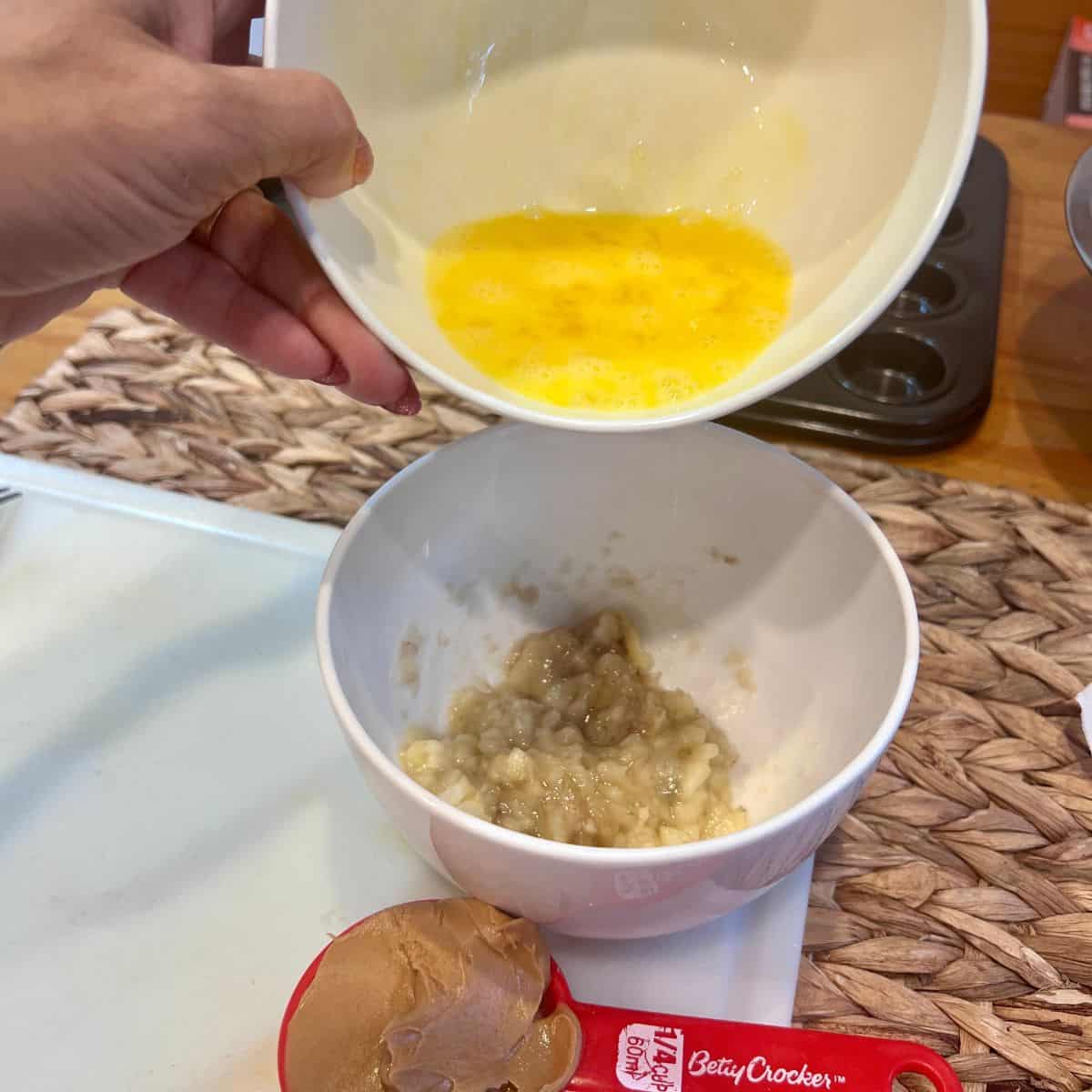 Hand pouring a beaten egg from a small bowl into mashed banana to make 3-ingredient peanut butter banana mini muffins.