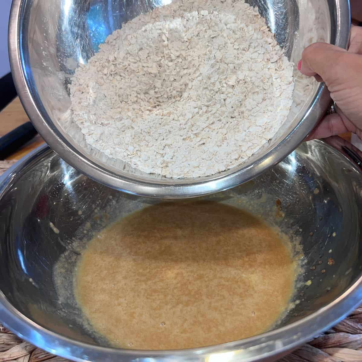 Hand pouring a bowl of dry oatmeal and flour ingredients into a mixing bowl of wet ingredients to make oatmeal cream pie cookie dough.