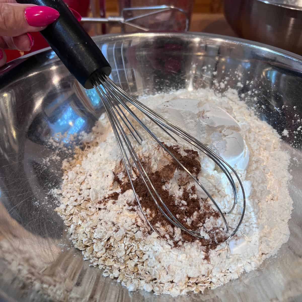 Hand whisking rolled oats, flour, cinnamon, and other dry ingredients in a mixing bowl for homemade oatmeal cream pie cookies.