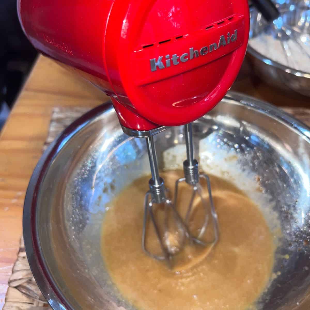 Red KitchenAid cordless hand mixer blending wet ingredients in a mixing bowl for homemade oatmeal cream pie cookies.