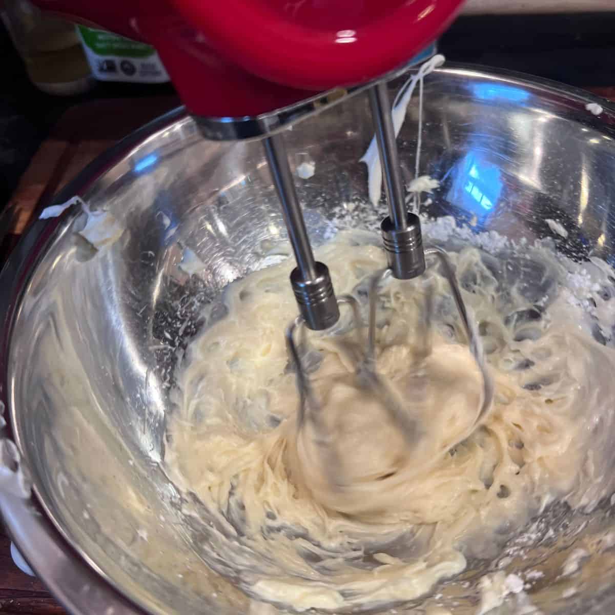 Red cordless hand mixer blending creamy marshmallow filling in a mixing bowl for homemade oatmeal cream pie cookies.