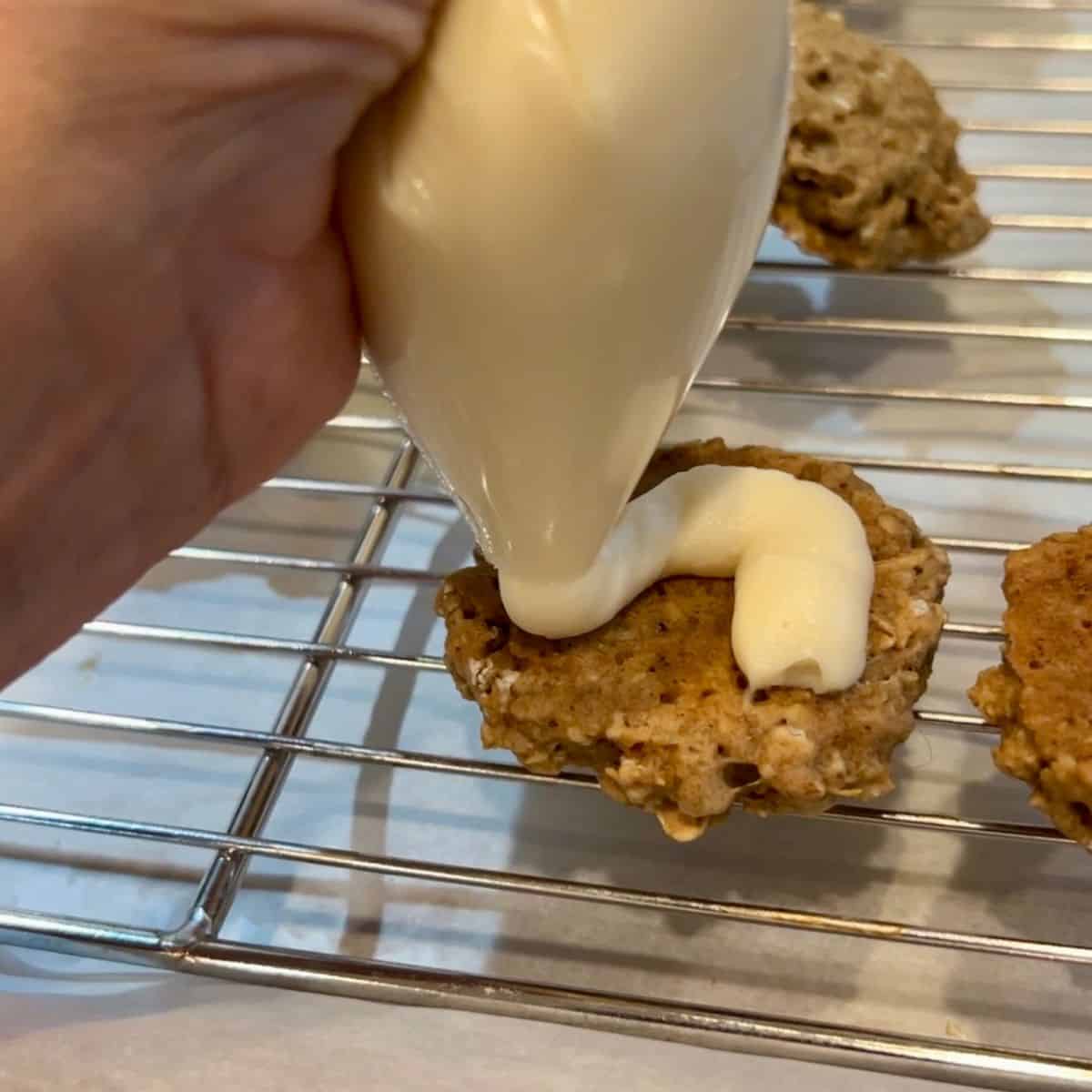 Hand squeezing marshmallow cream filling from a plastic bag with the corner snipped onto a homemade oatmeal cookie.